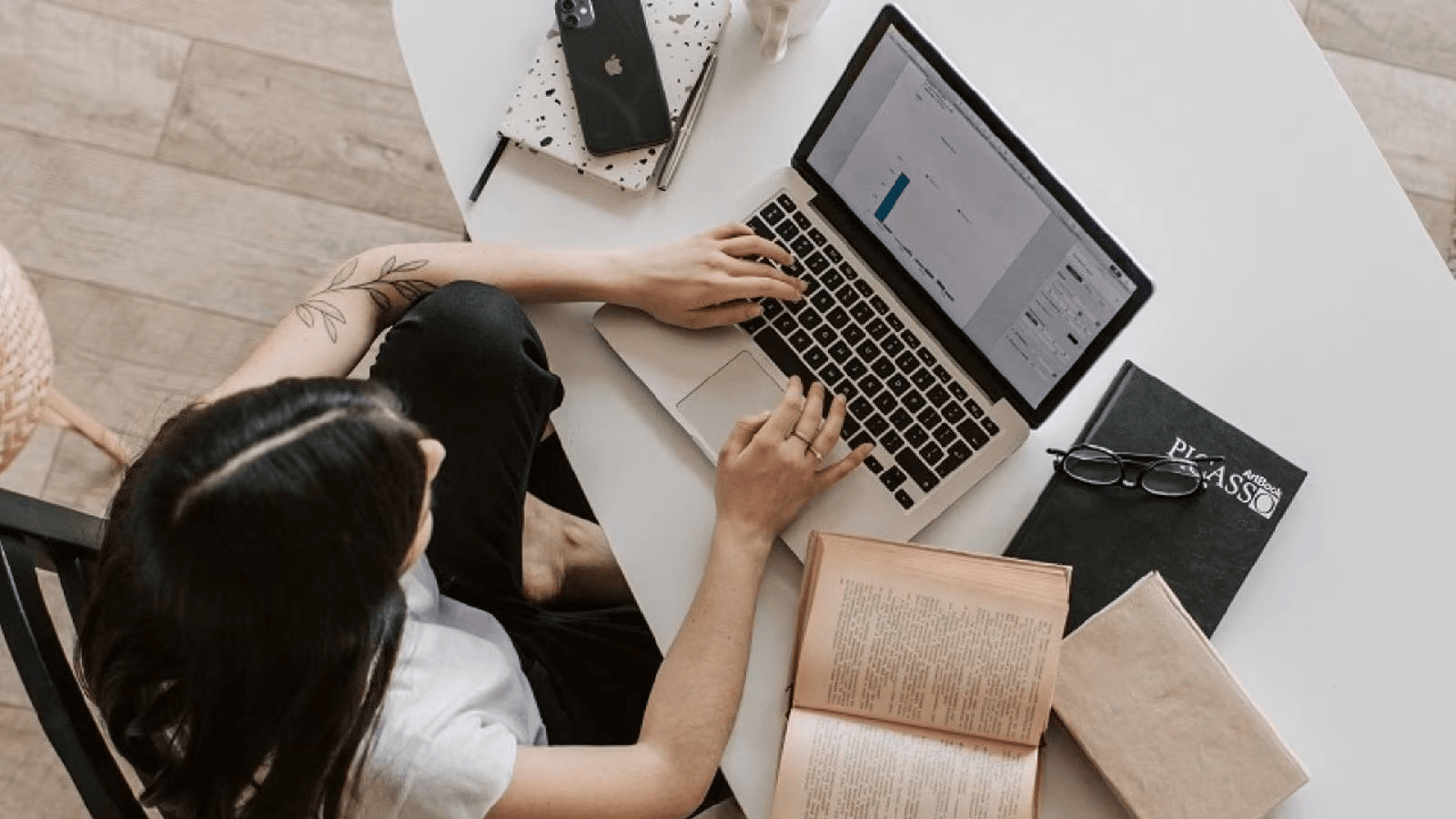 A woman sits at a table with a laptop and books, focused on her work in a cozy study environment.