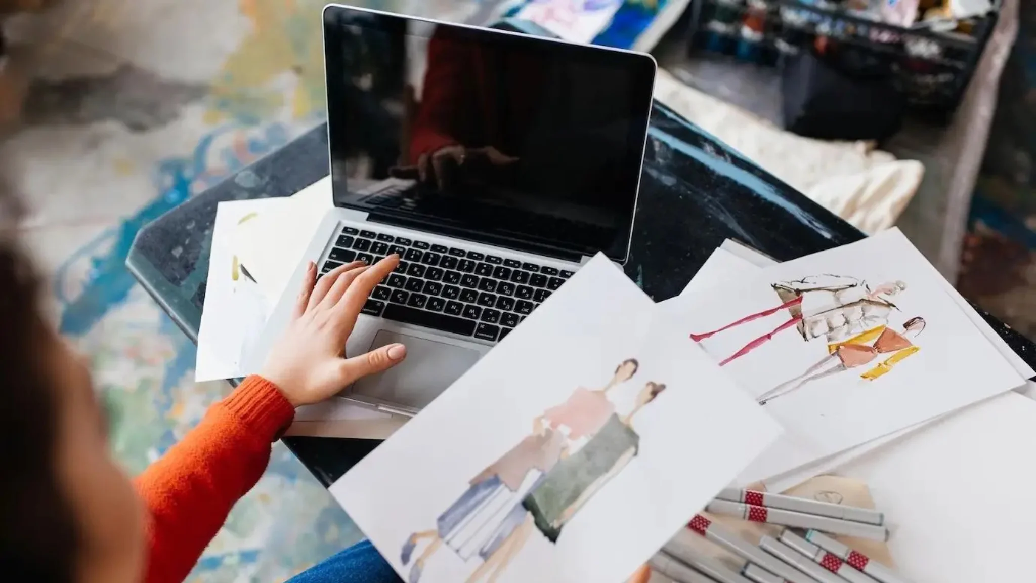 A woman works on her laptop while holding a sketchbook, blending digital and traditional creativity.