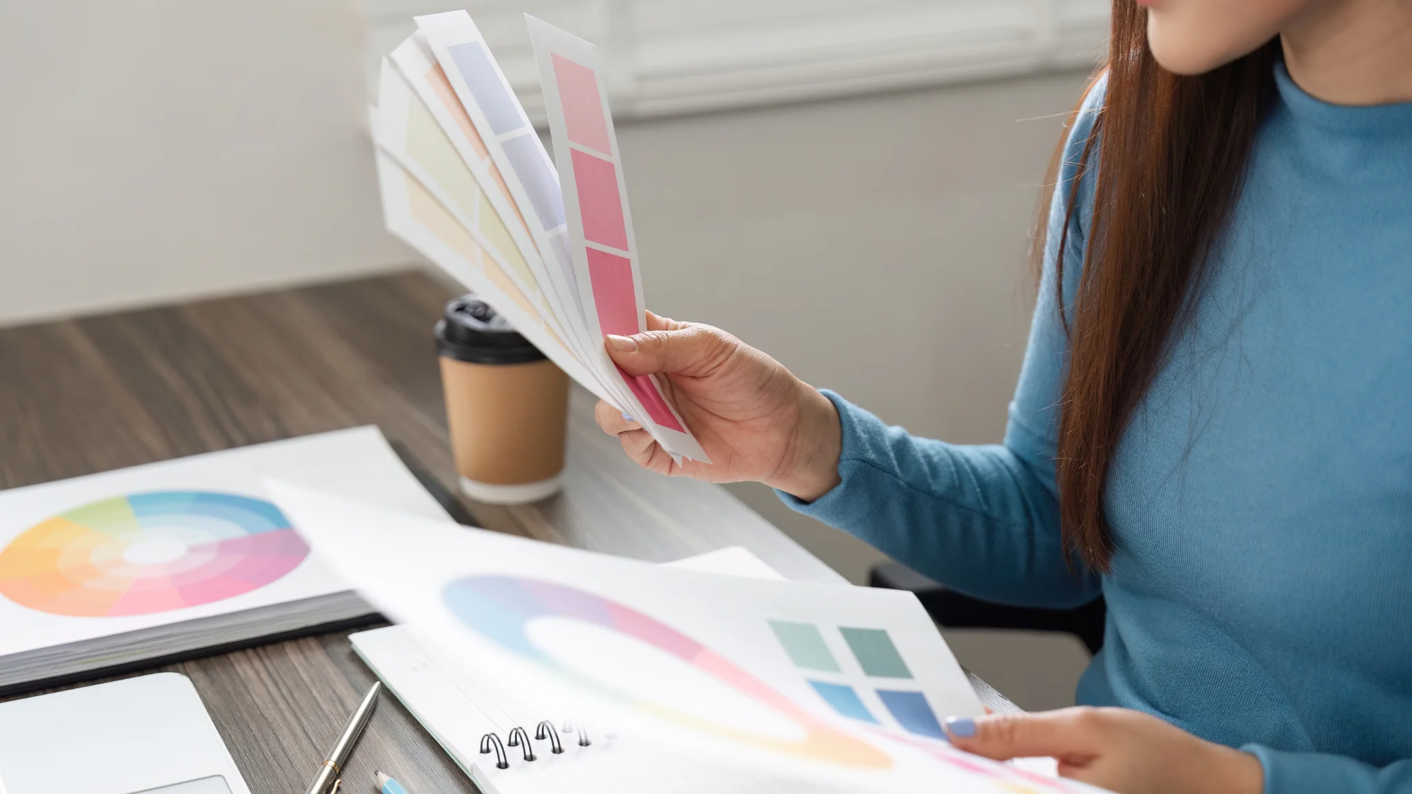 A woman sitting at a desk holds a color swatch, contemplating her design choices with a thoughtful expression.