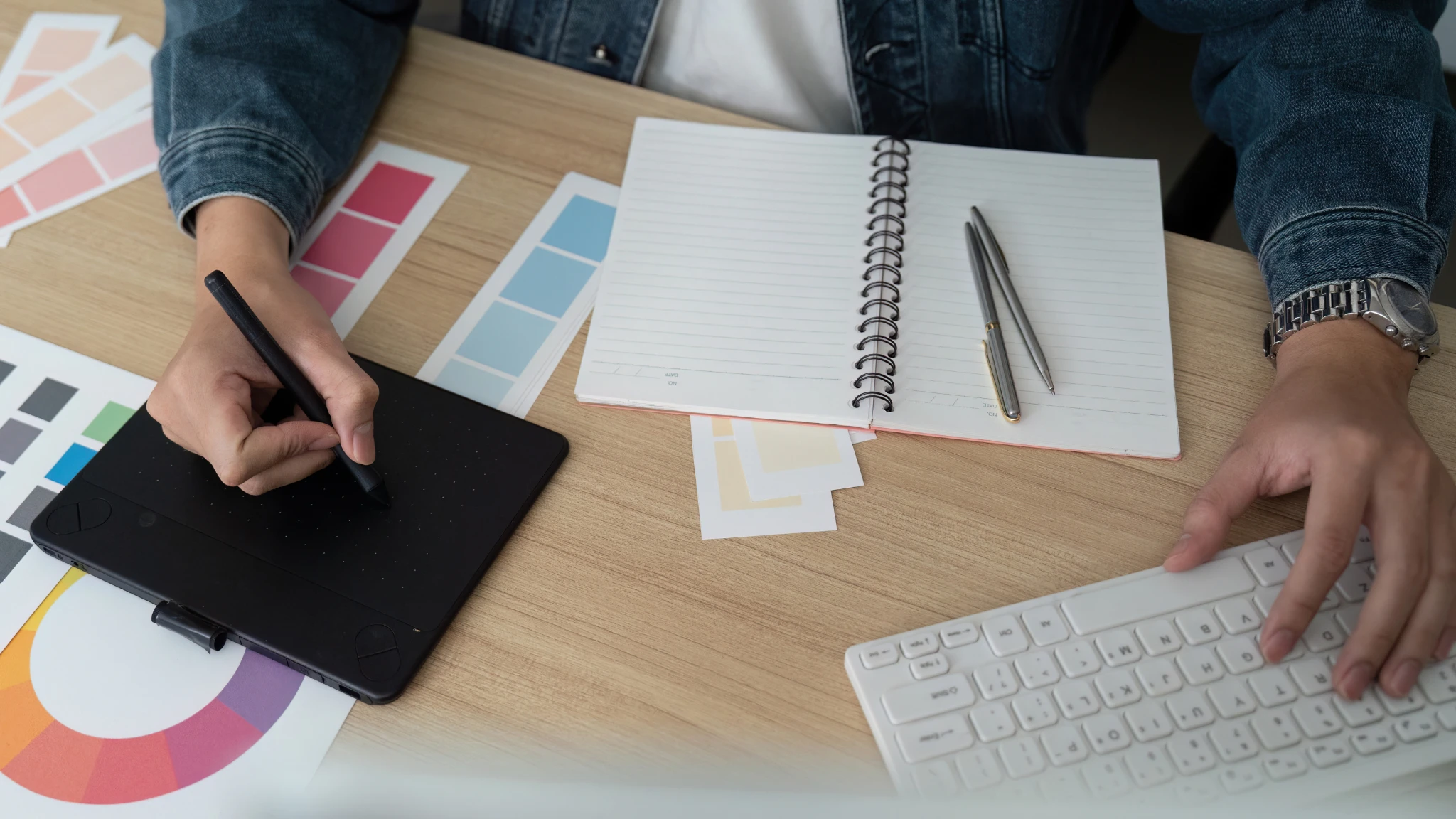 A person types on a computer while holding a pen and jotting notes in a notebook beside them.