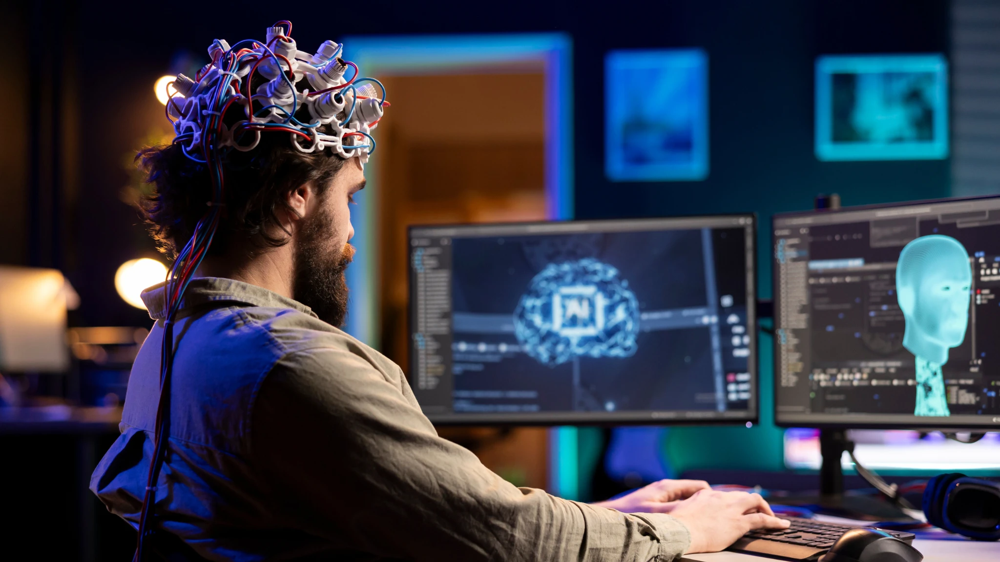 A man in a hat sits at a computer, focused on the screen in a cozy workspace.