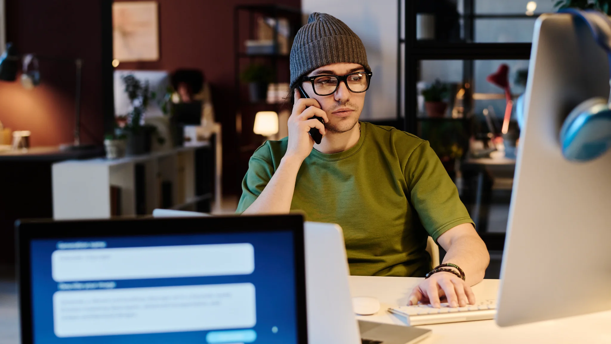 A man wearing a beanie sits at a desk, talking on the phone with a focused expression.