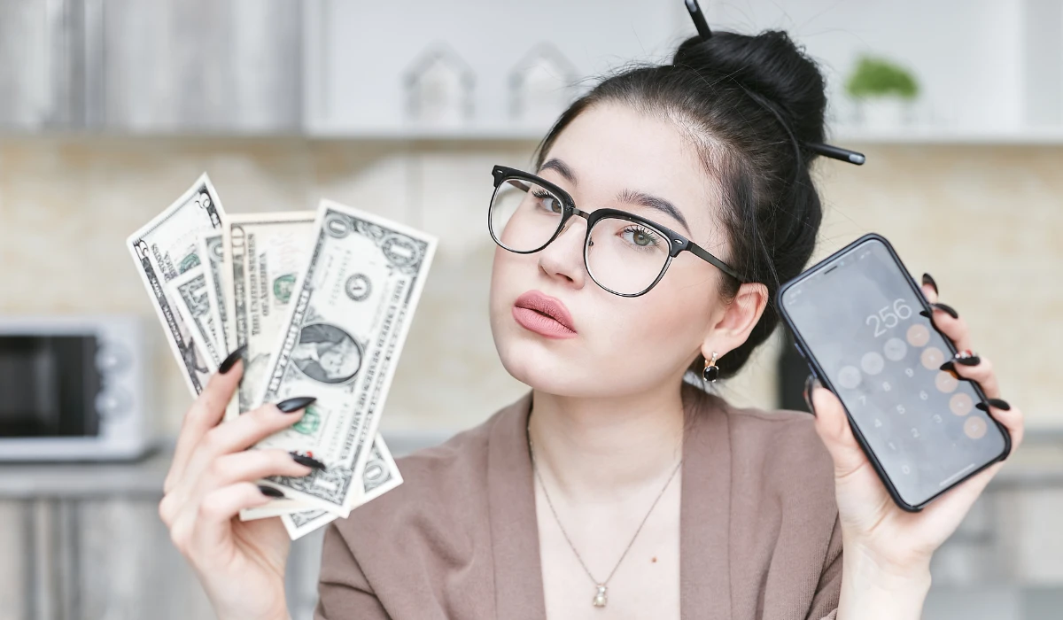 A woman smiling while holding cash in one hand and a smartphone in the other.