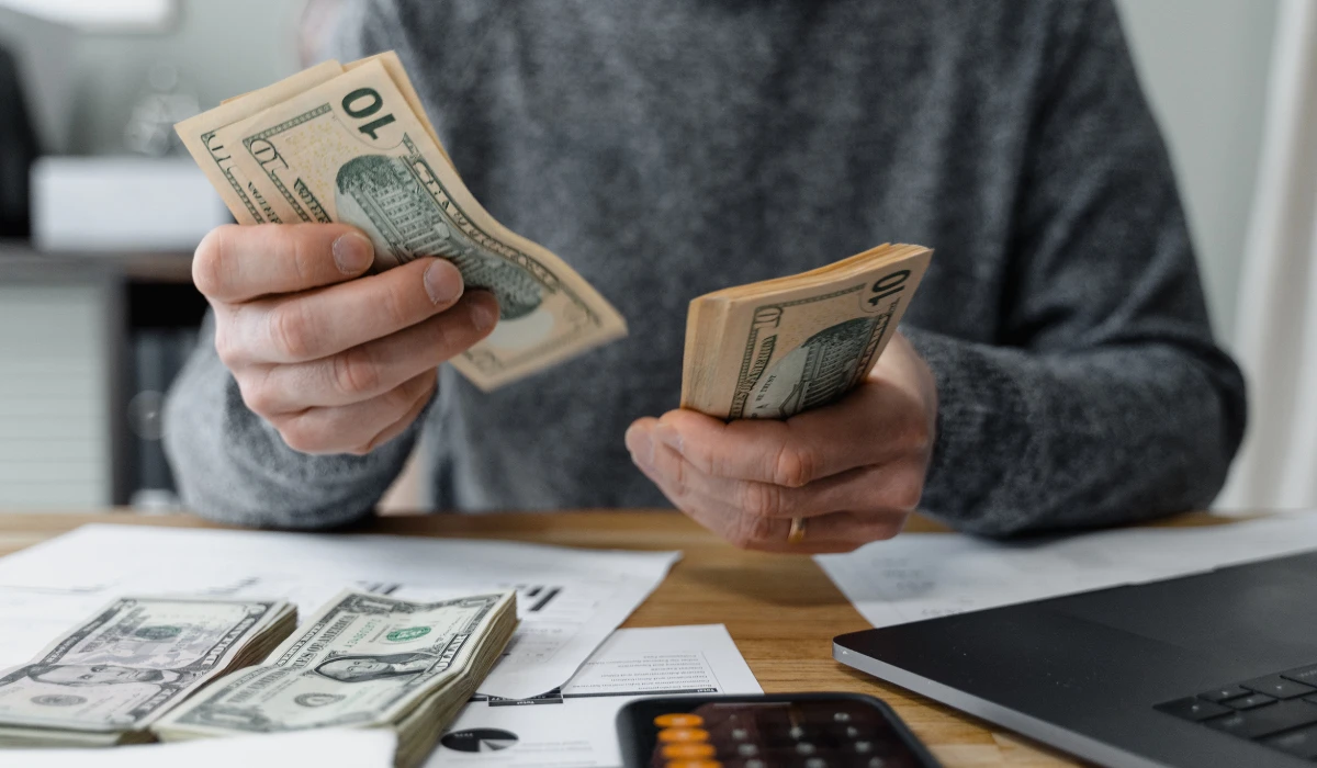A man smiles while holding cash in front of a laptop, suggesting online shopping or financial planning.