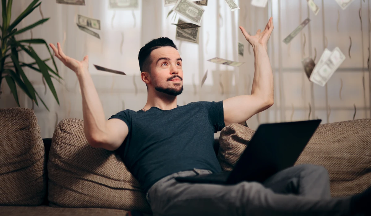 A man sits on a couch with a laptop as money falls from the ceiling around him, creating a whimsical scene.