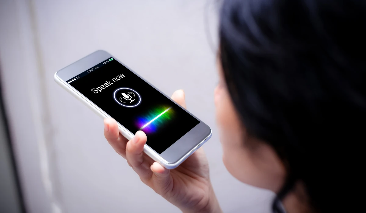 A woman smiling while holding a smartphone displaying a voice assistant interface.