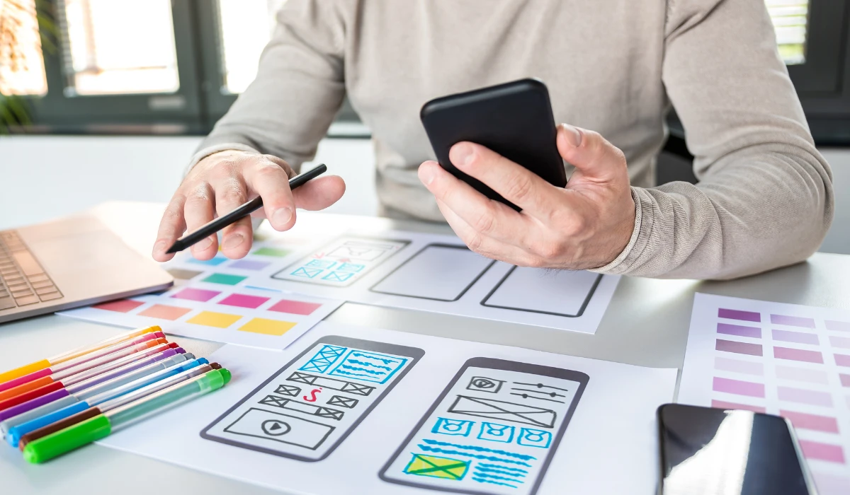 A man designs a website using a smartphone in one hand and a laptop on the table in front of him.