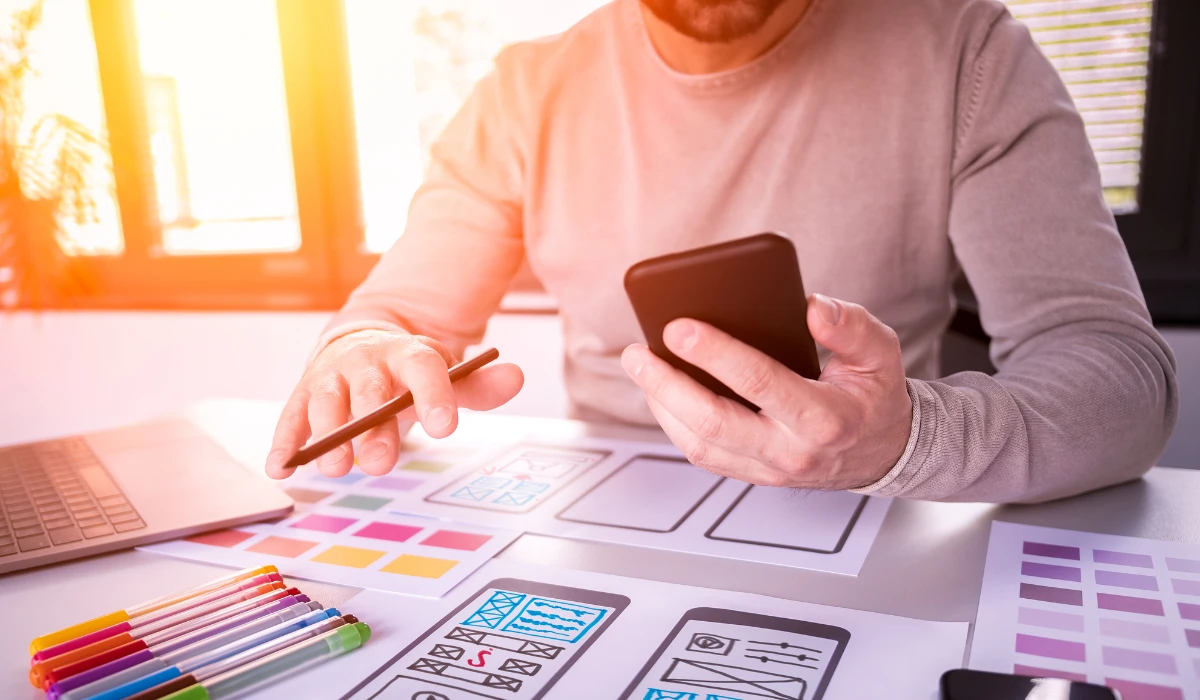 A man sits at a desk using his phone, surrounded by a laptop and various design tools.