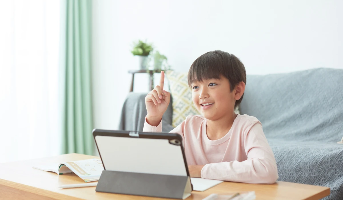 A boy sits at a table, focused on his laptop, with a bright smile on his face.