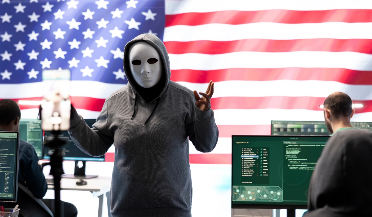 A man in a mask holds a computer in front of an American flag, symbolizing technology and patriotism.