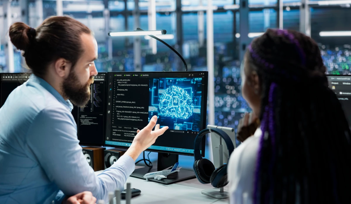 Two people collaborate at computers, focused on a large screen displaying data or visuals.