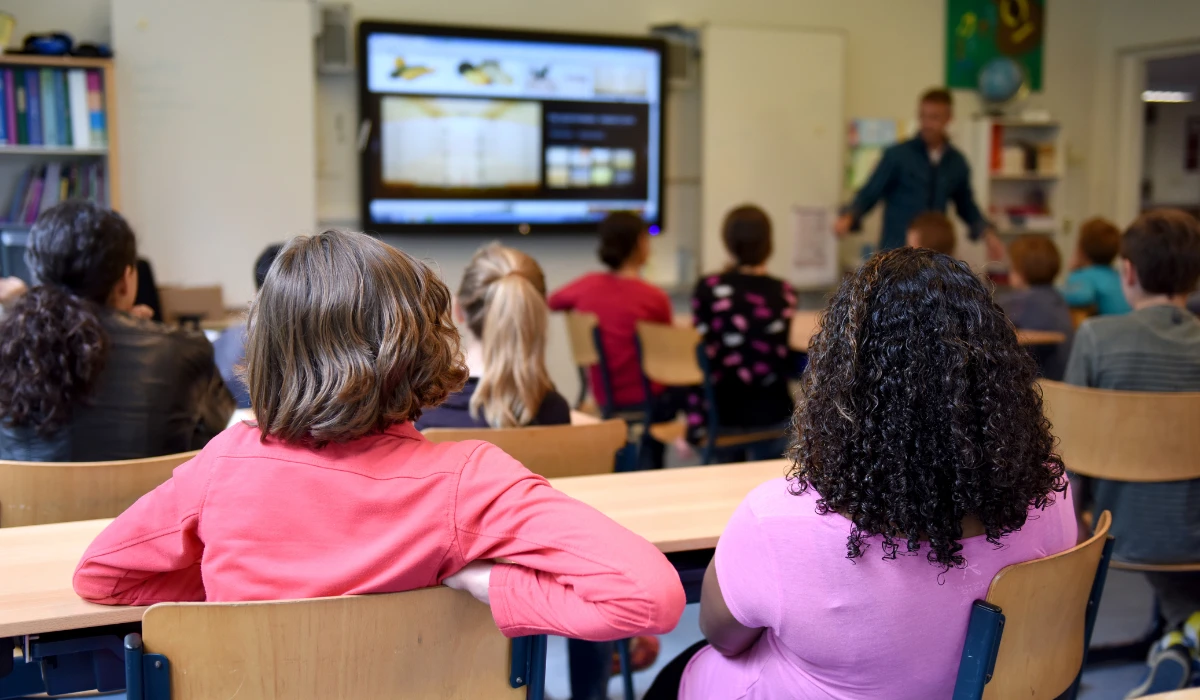A diverse group of children in a classroom, all eyes on a video playing on the screen, showing interest and excitement.