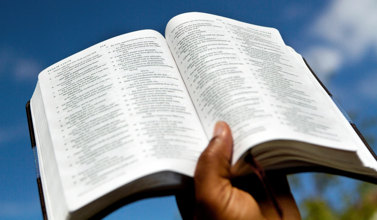 A person holds an open Bible against a bright blue sky, symbolizing faith and inspiration.