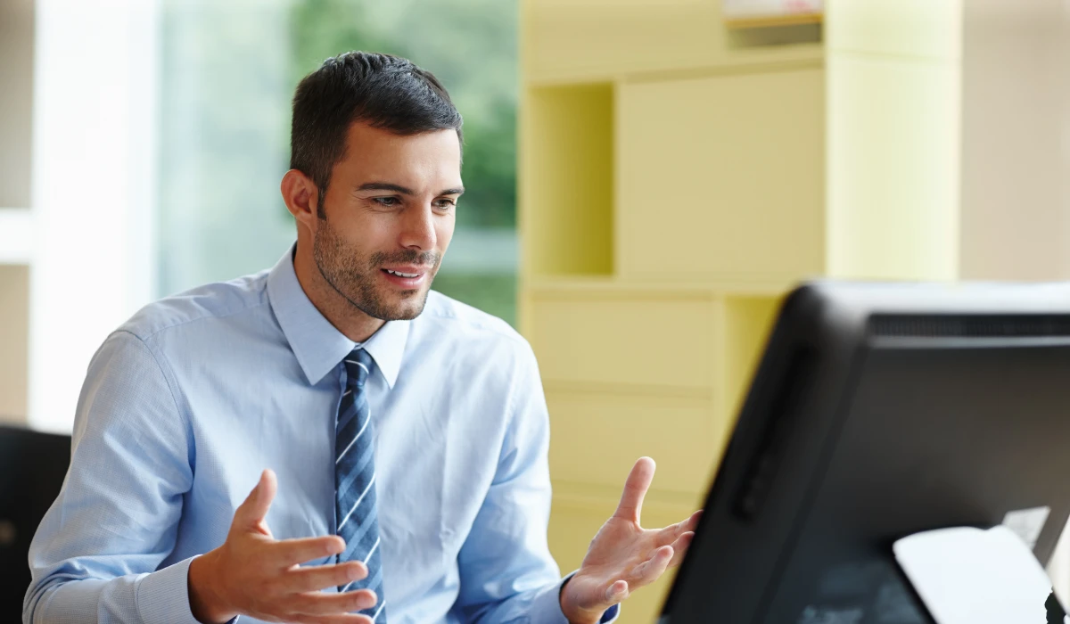A man wearing a blue shirt and tie is seated at a desk with a computer in front of him.